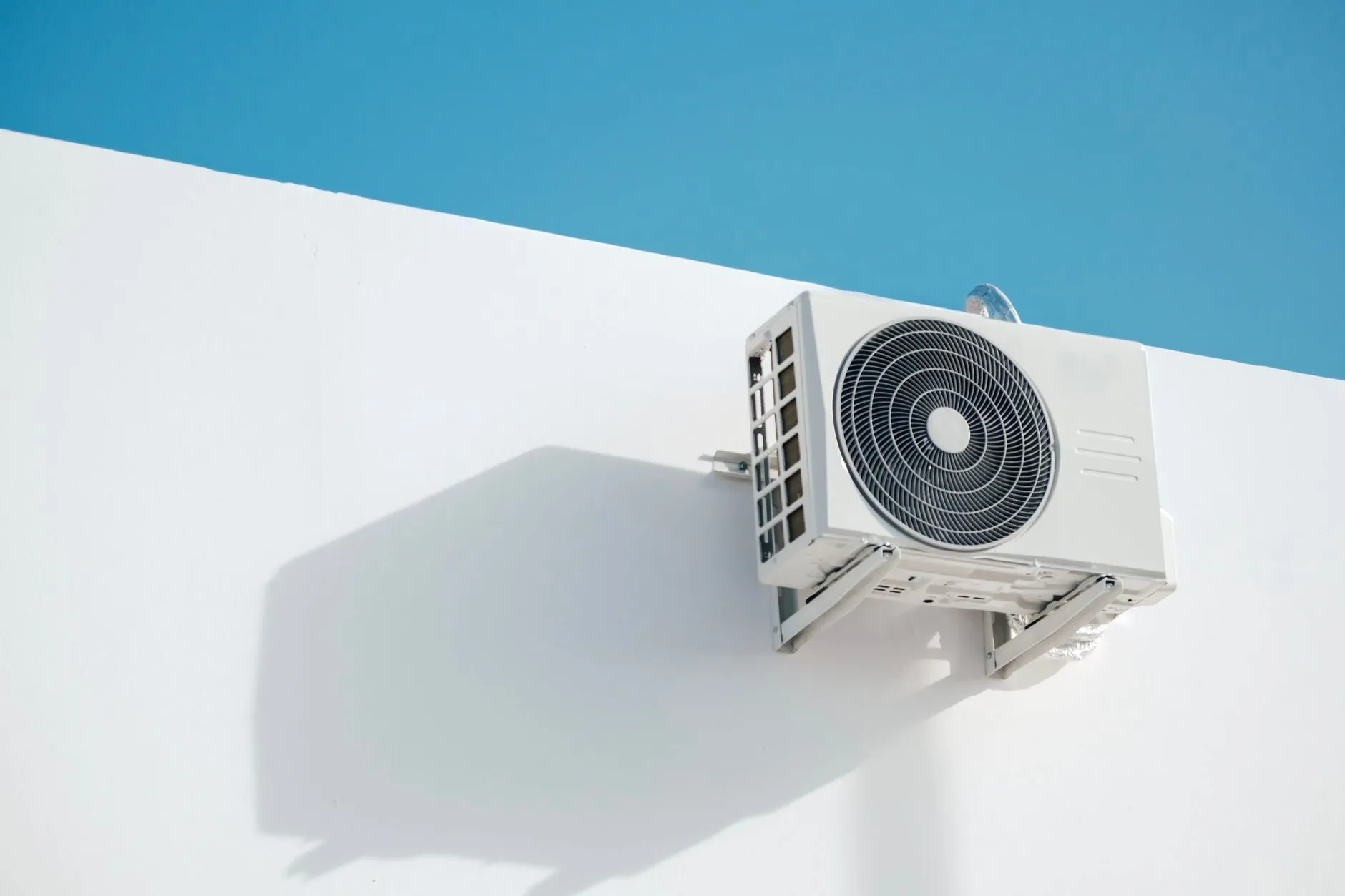A white outdoor air conditioning unit on brackets high on a white rendered wall, against a blue sky