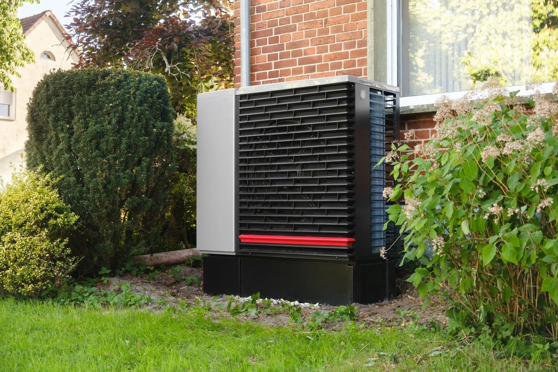 An air source heat pump on a plinth beside a red-brick house, between garden shrubs