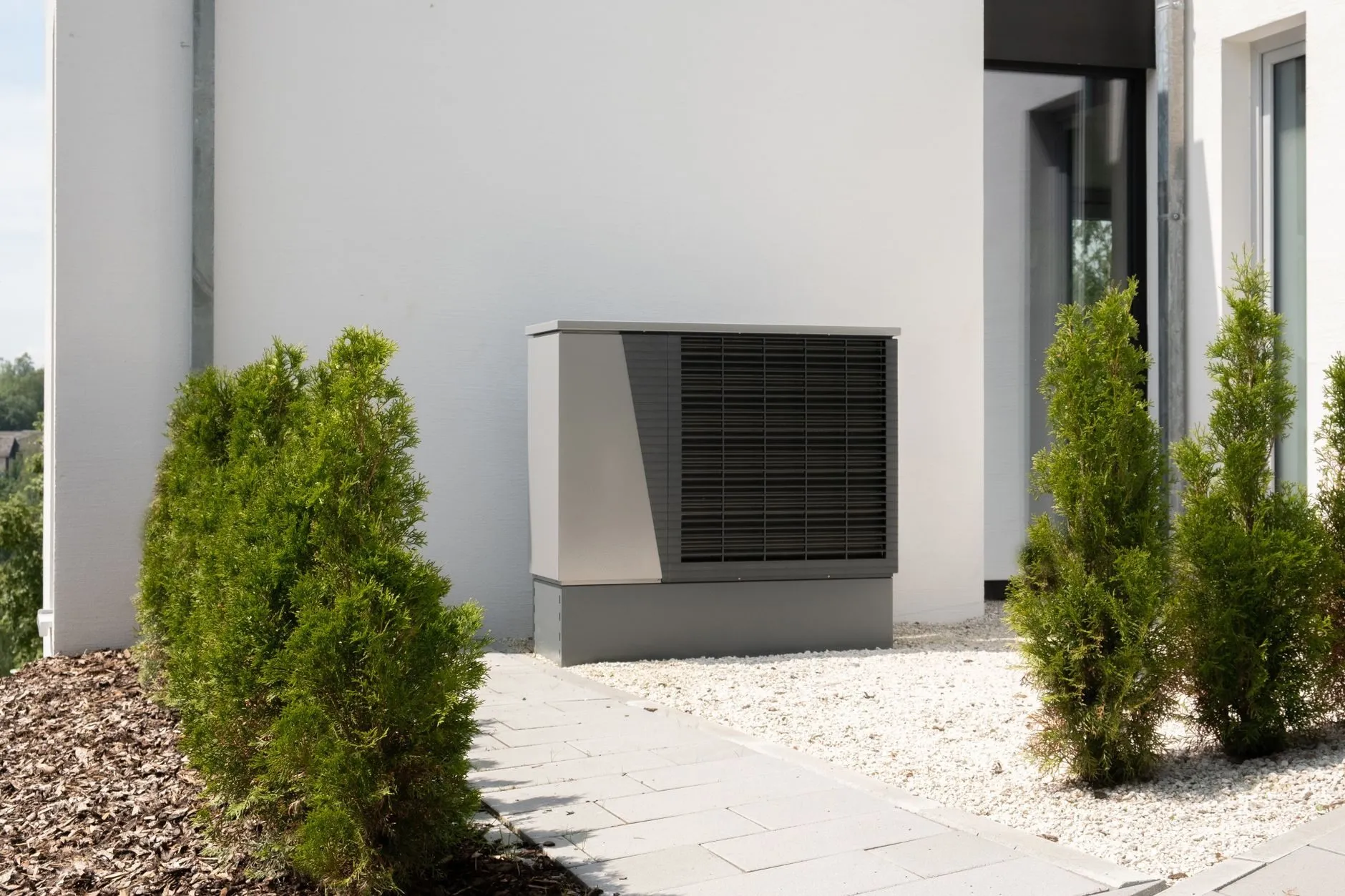 A heat pump beside a paved garden path between conifers